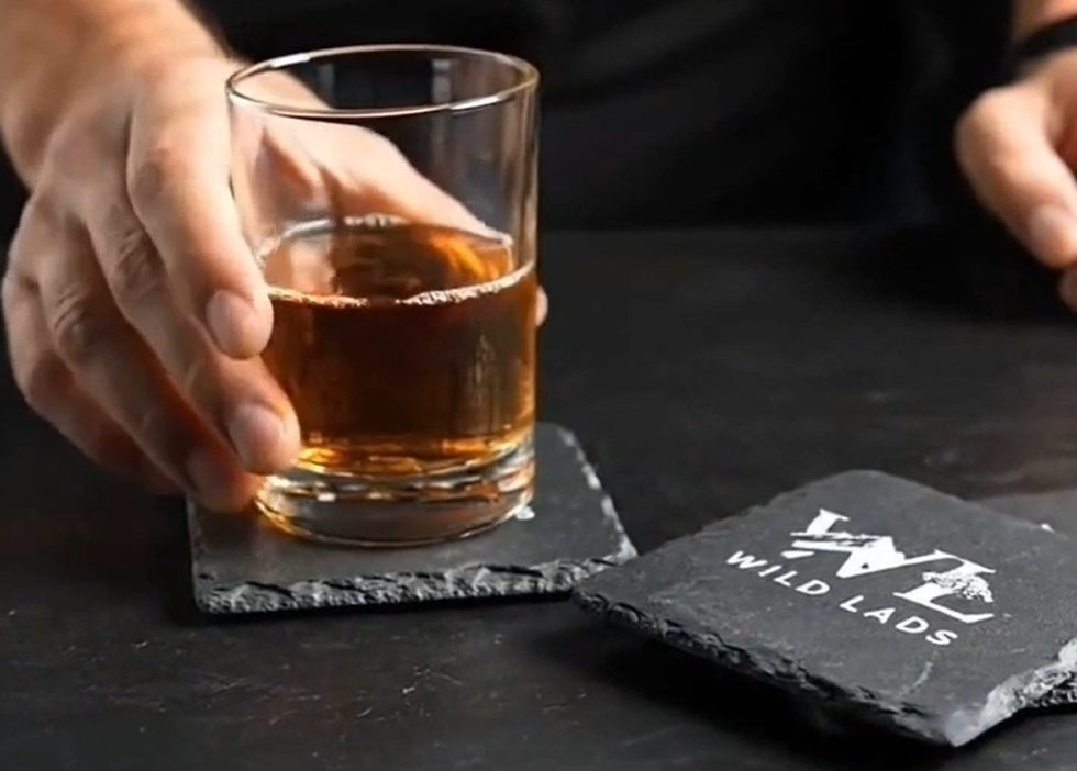 Hand holding a glass of whiskey on a slate coaster with 'Wild Lads' branding.